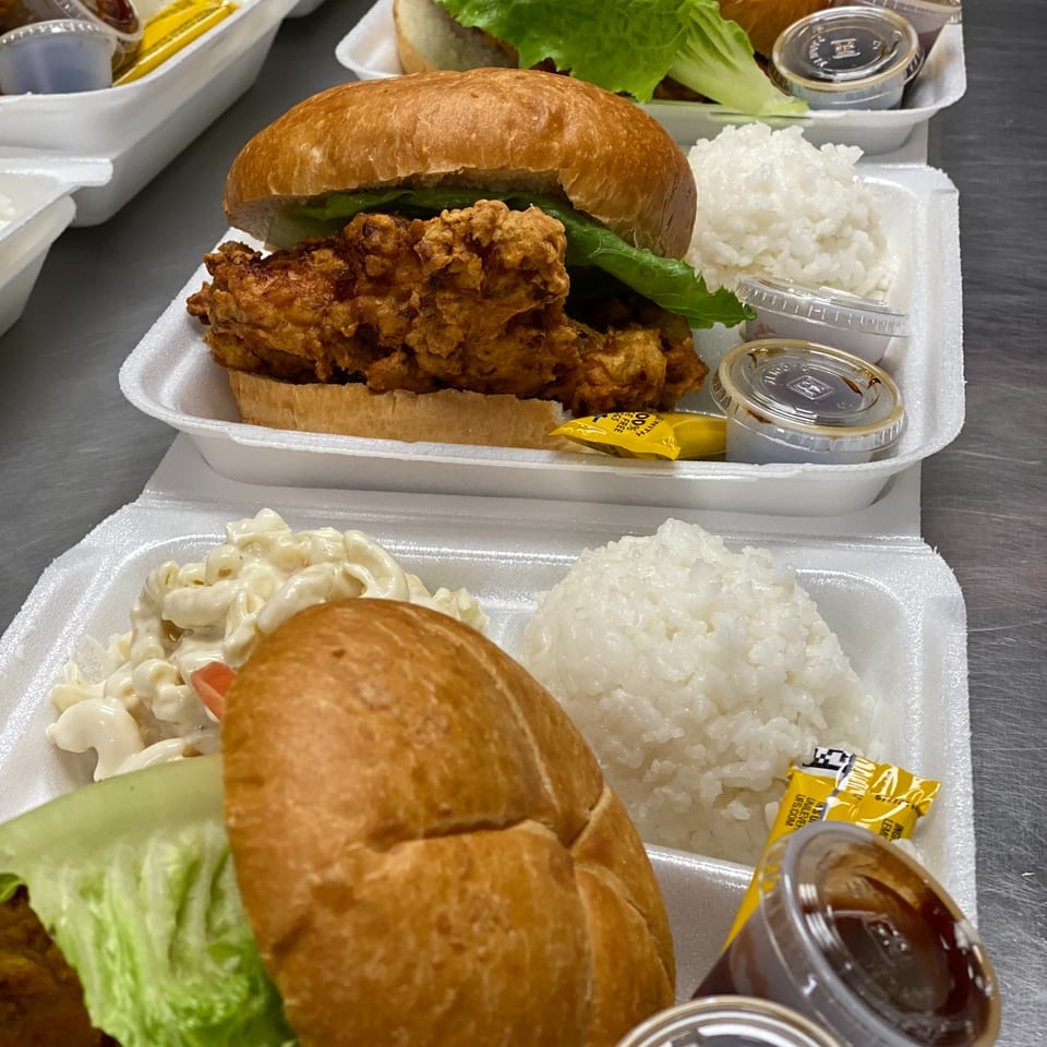 Crispy fried chicken sandwiches with lettuce, white rice, macaroni salad, sauces, and condiments in takeout containers.