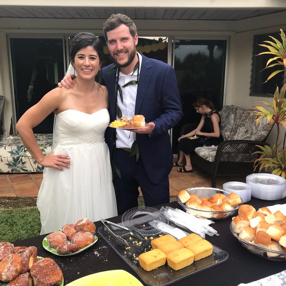 Newlywed couple pose beside a buffet table filled with rolls, pastries, cornbread, and other desserts at an outdoor reception.