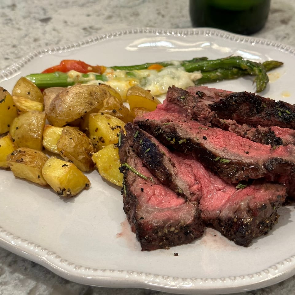 Sliced medium-rare steak with roasted potatoes, asparagus, tomatoes, and creamy vegetables on a white plate.