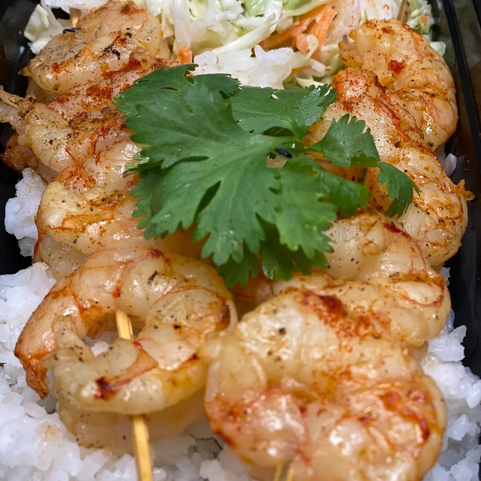 Grilled seasoned shrimp skewers served over white rice with mixed salad and fresh cilantro in a black takeout tray.