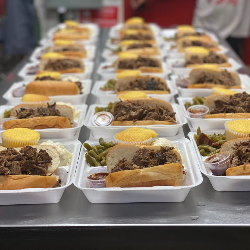 Rows of takeout meals featuring meat sandwiches, green beans, dipping sauces, and yellow cupcakes in foam containers.