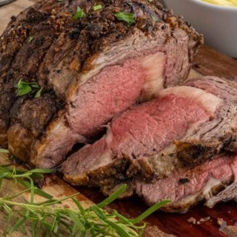 Sliced medium-rare roast beef with a seasoned crust rests on a wooden board alongside fresh herbs and creamy sauce.