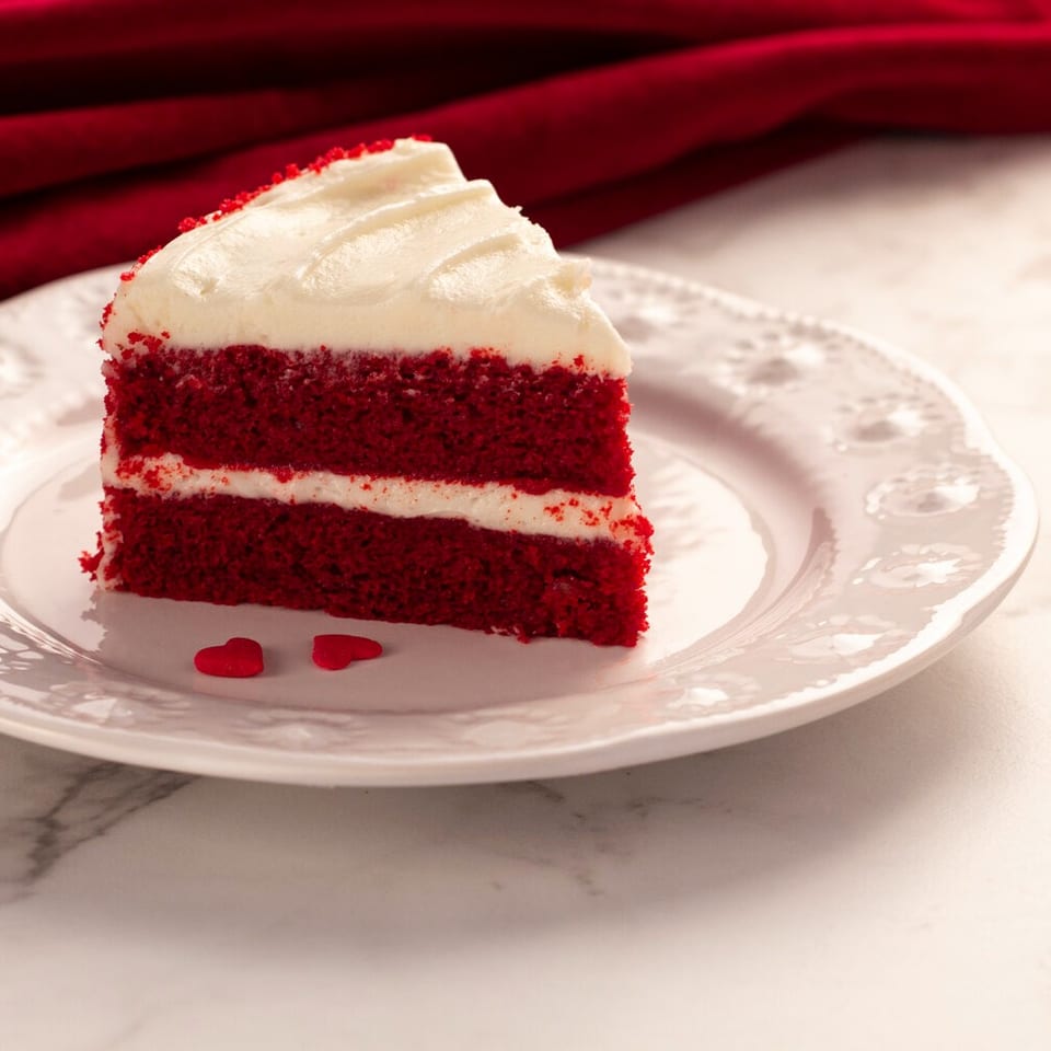 Slice of red velvet cake with cream cheese frosting on an ornate plate, garnished with red heart-shaped sprinkles.