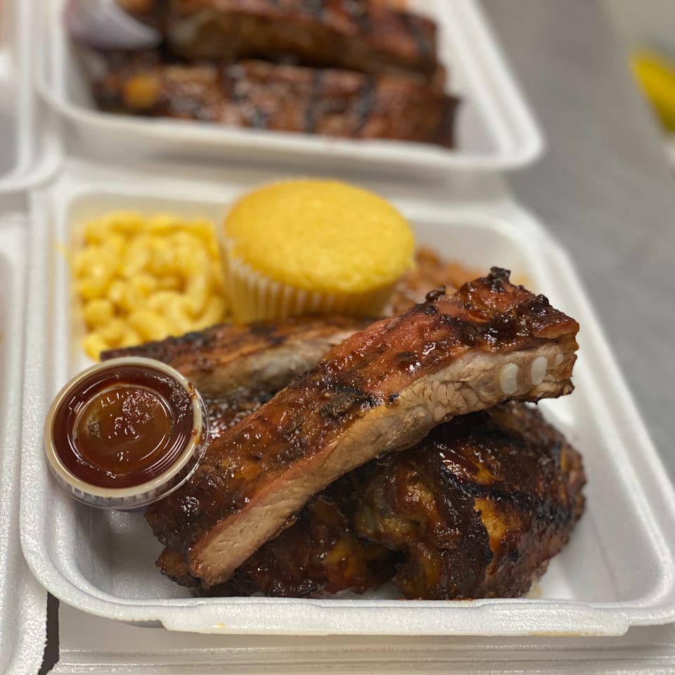 Barbecue ribs with sauce, macaroni and cheese, cornbread, and baked beans served in a takeout container.