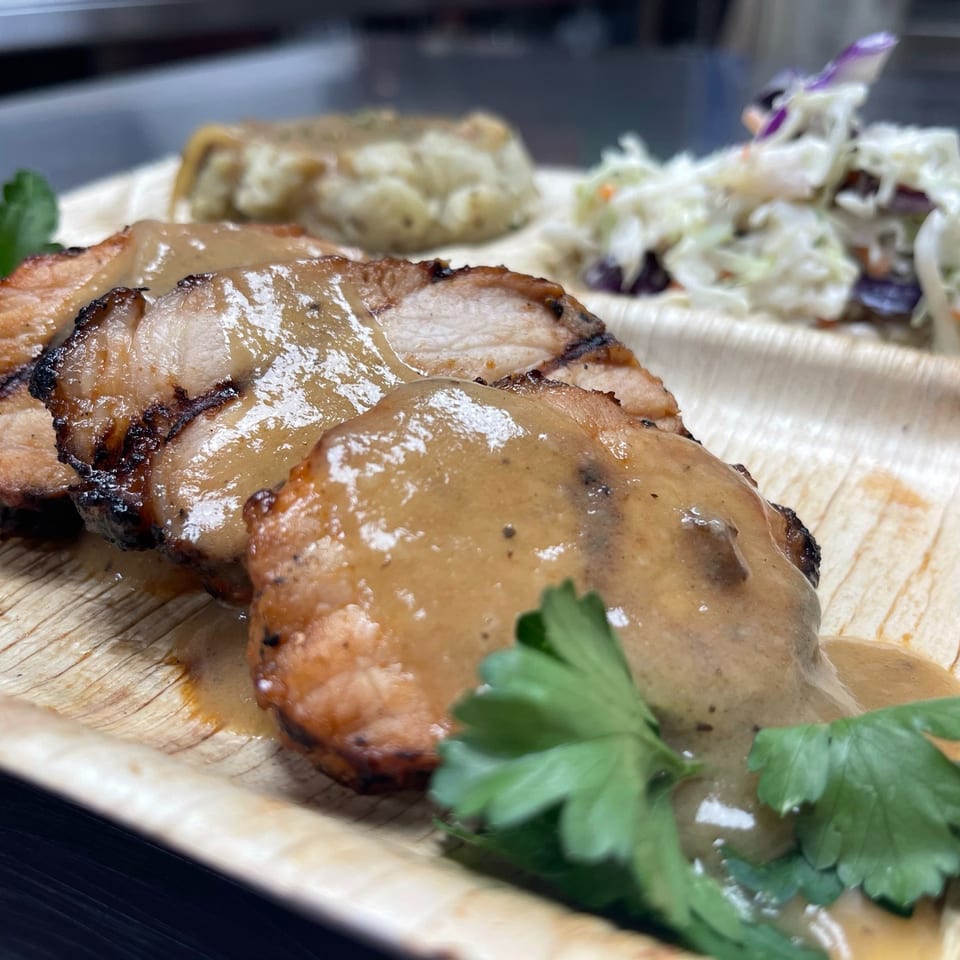 Grilled sliced meat topped with brown sauce, served on a wooden platter with coleslaw, vegetables, and parsley.