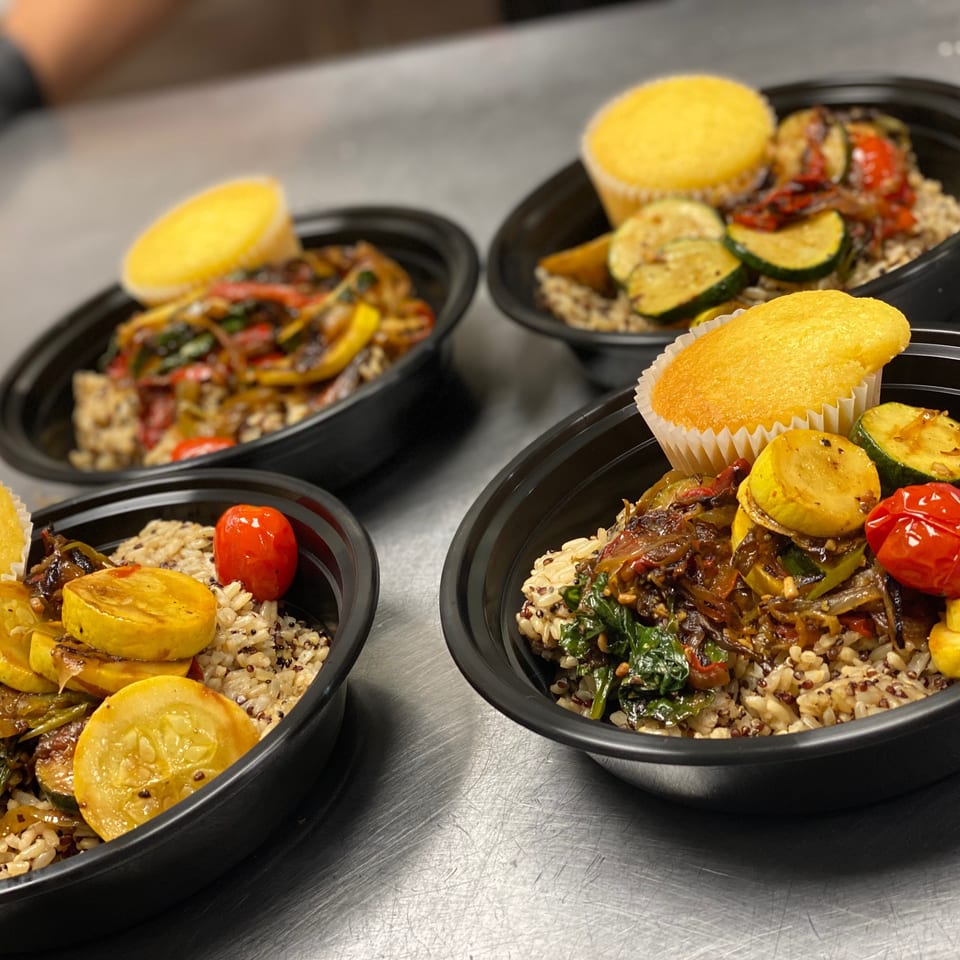 Black bowls of multigrain rice topped with roasted vegetables and golden cornbread muffins on a stainless-steel counter.