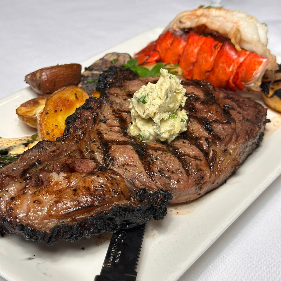 Grilled steak topped with herb butter, served with lobster tail, roasted vegetables, and a steak knife on a white plate.