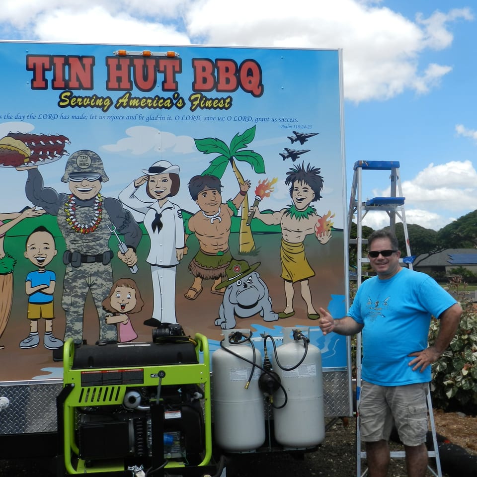 Tin Hut BBQ trailer featuring a colorful multicultural mural, with a man standing beside propane tanks and cooking equipment.
