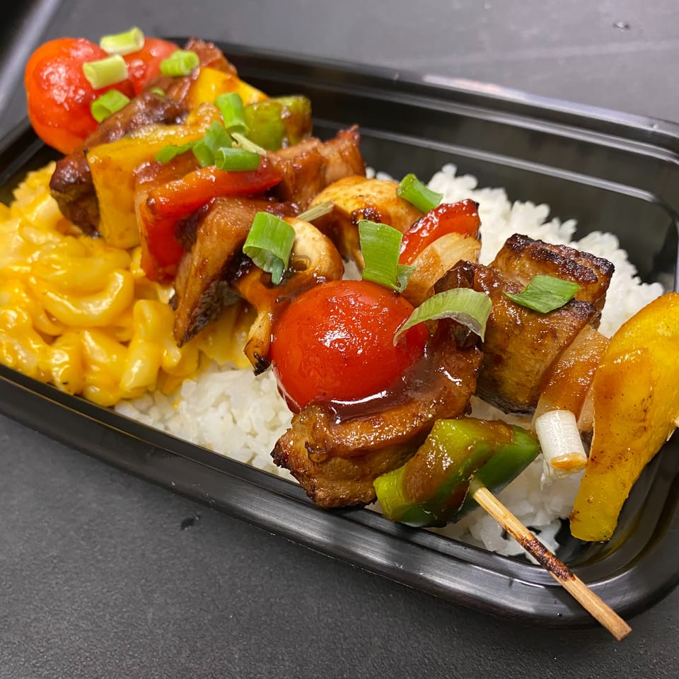 Grilled meat and vegetable skewers with white rice and macaroni and cheese in a black takeout tray.