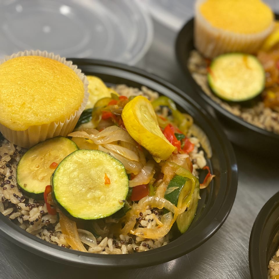 Meal-prep bowls feature quinoa, sautéed zucchini and peppers, and yellow cornbread muffins in black containers.