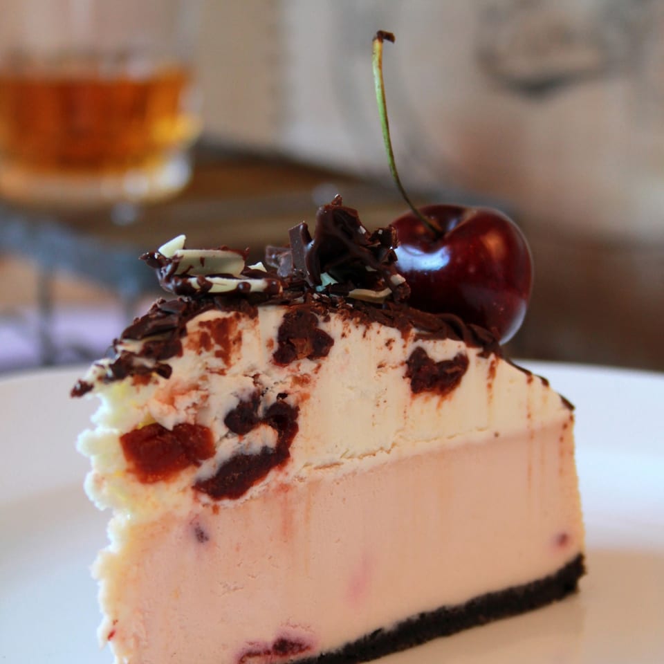 Slice of cherry ice cream cake with a chocolate cookie crust, chocolate shavings, nuts, and a cherry garnish.