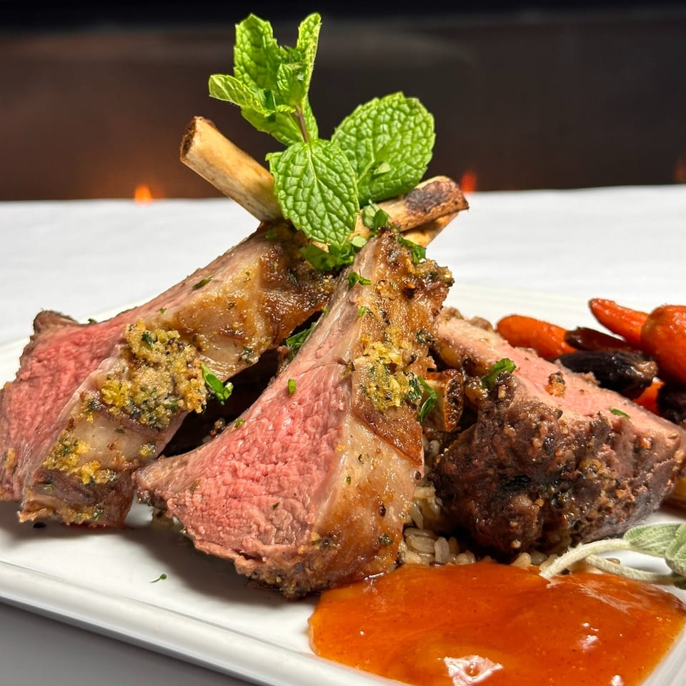 Sliced medium-rare lamb chops with mint garnish, roasted vegetables, and orange sauce on a white plate.