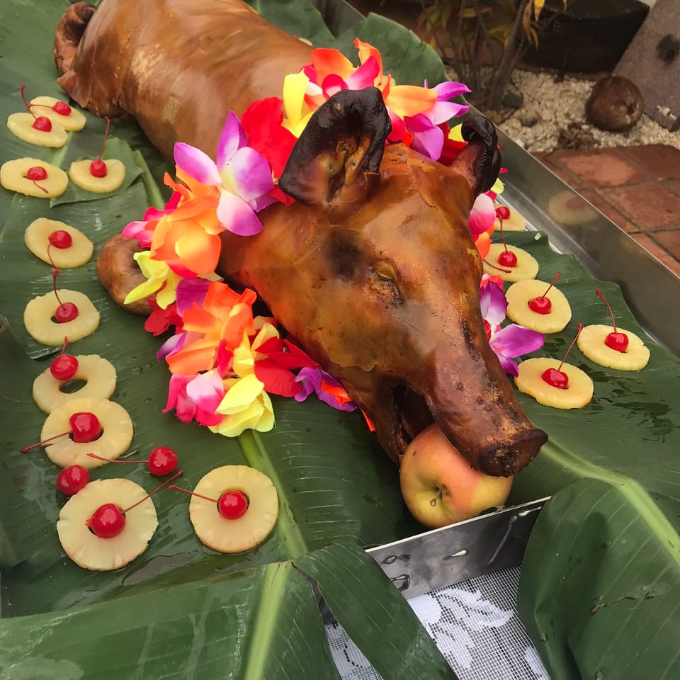 Whole roasted pig displayed on banana leaves, surrounded by tropical flowers, pineapple slices, cherries, and fruit.