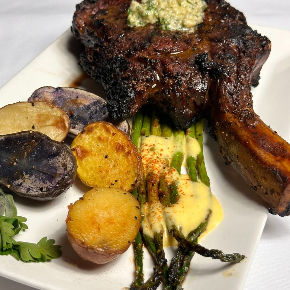 Grilled bone-in steak topped with herb butter, served with roasted potatoes and asparagus drizzled with creamy sauce.