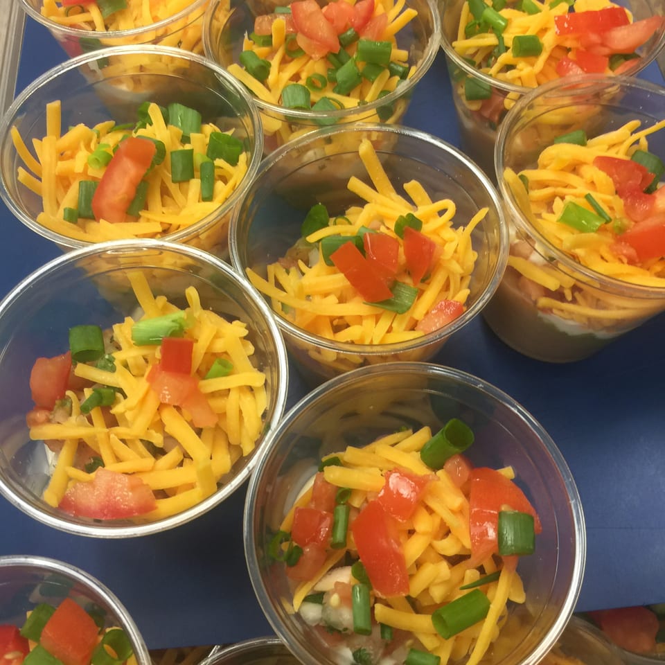Clear cups filled with shredded cheddar cheese, diced tomatoes, and chopped green onions arranged on a blue surface.