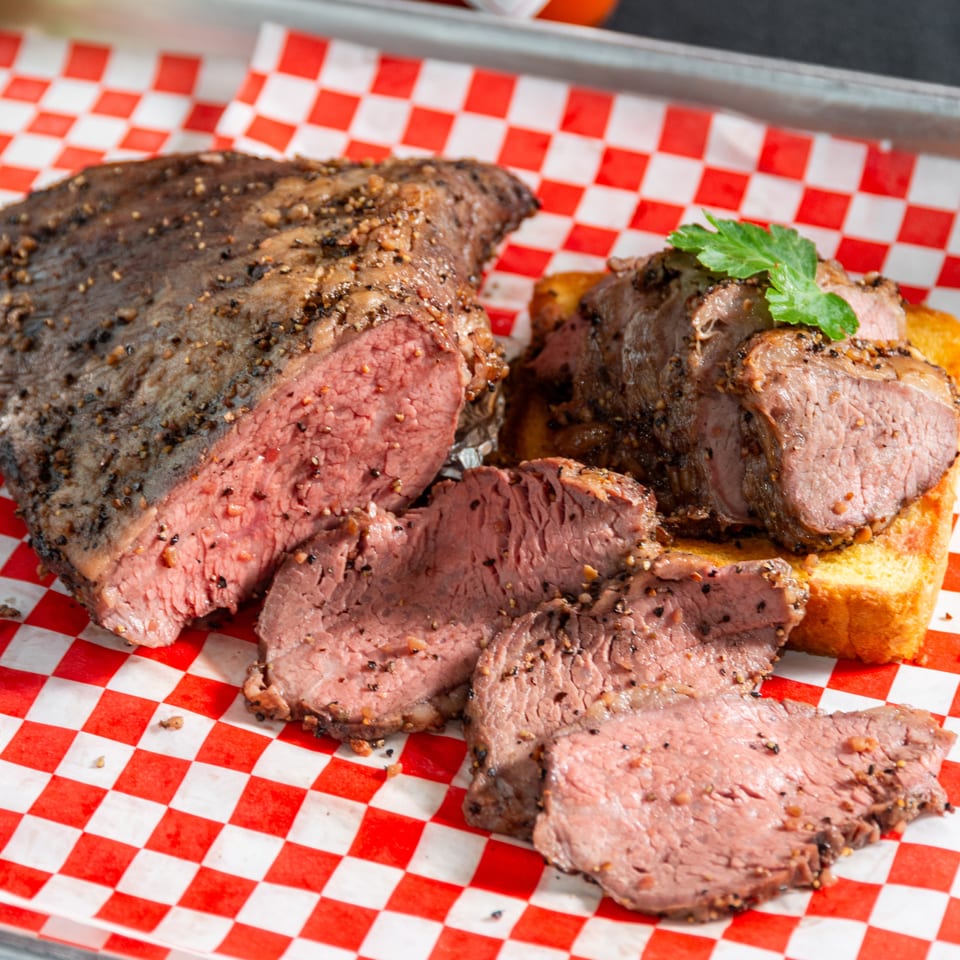 Pepper-crusted medium-rare roast beef slices served with a browned bread roll on red-and-white checkered paper.