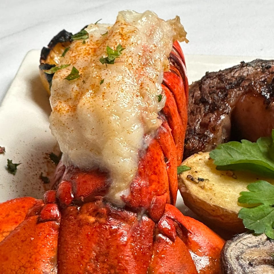 Close-up of a seafood platter featuring lobster, shrimp, grilled steak, roasted vegetables, and herbs on a white plate.