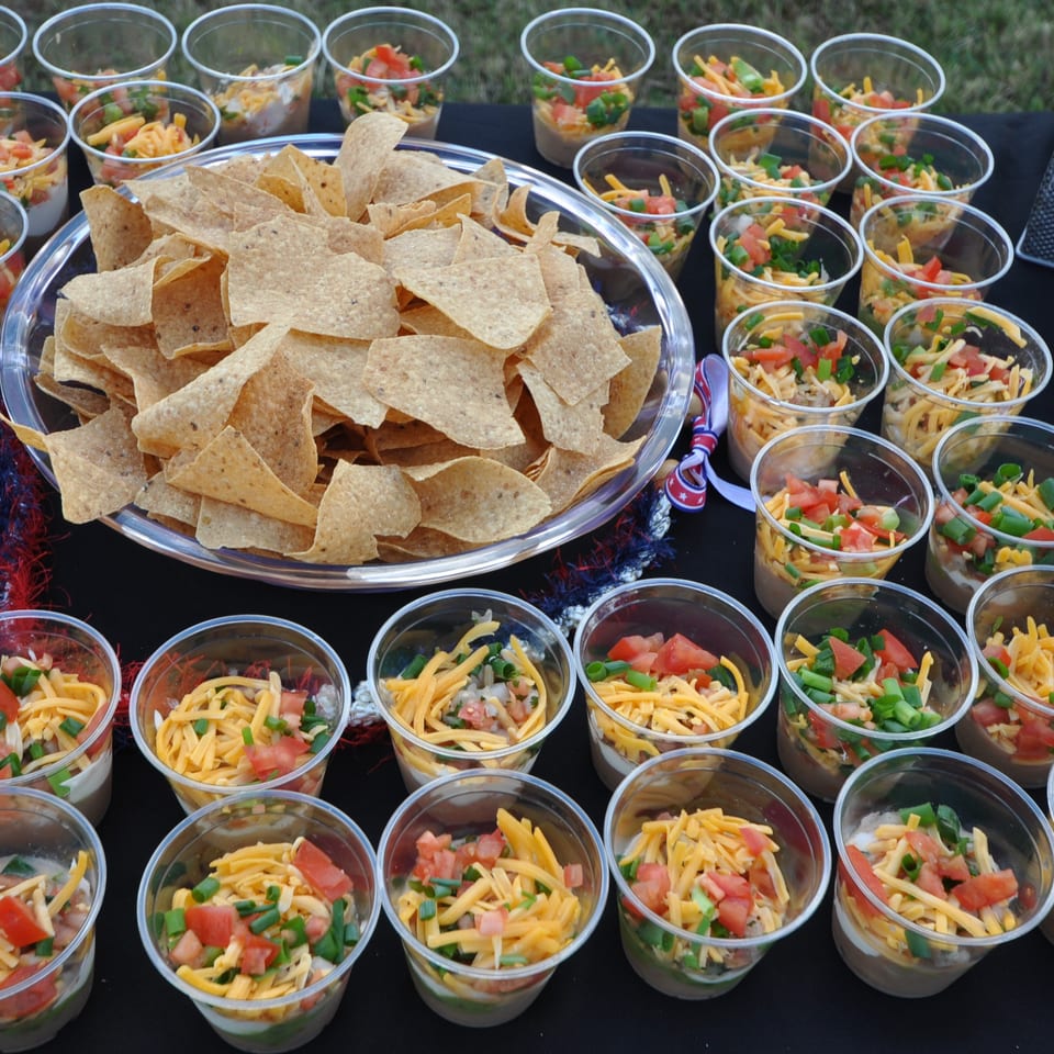 Large platter of tortilla chips surrounded by individual cups of layered dip topped with shredded cheese, tomatoes, and green onions.