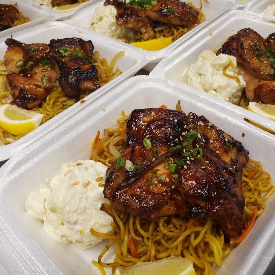 Takeout containers filled with glazed grilled chicken, chow mein noodles, creamy macaroni salad, lemon wedges, scallions, and sesame seeds.