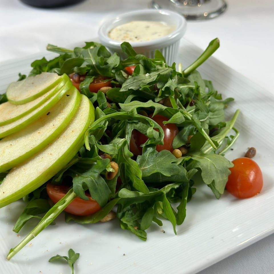Arugula salad with sliced green apple, cherry tomatoes, nuts, and creamy dressing on a white plate beside wine.