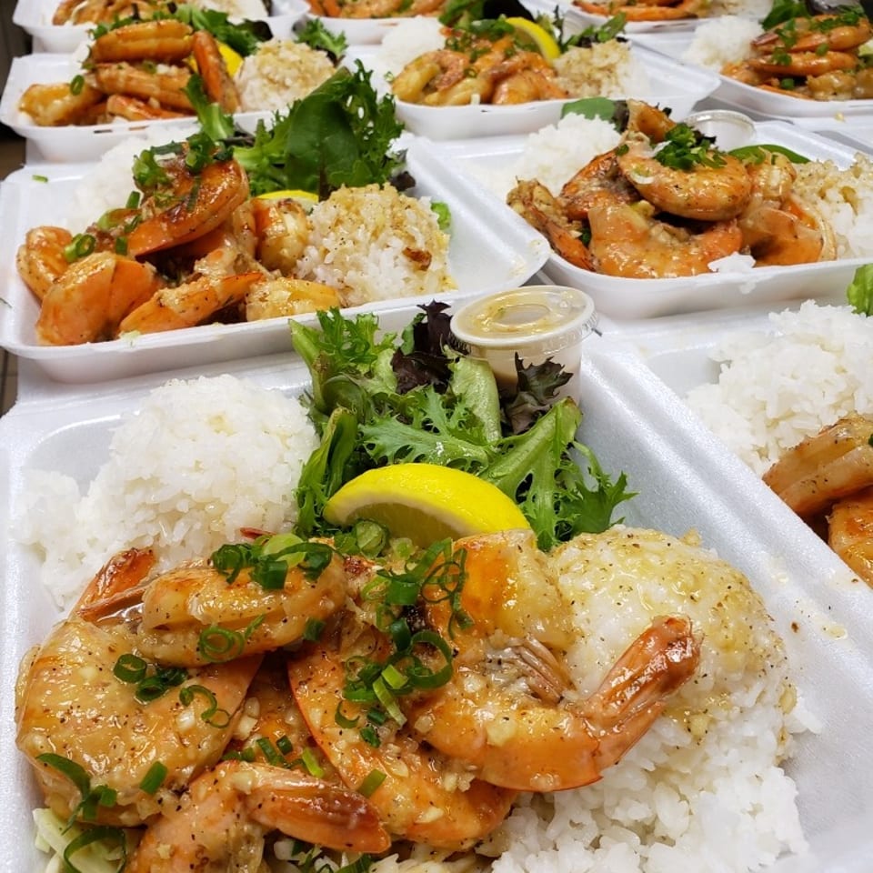 Rows of takeout trays filled with seasoned shrimp, white rice, greens, and lemon wedges.
