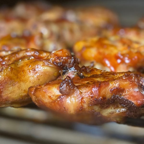 Close-up of Tin Hut BBQ chicken wings, glazed with sauce and char marks, cooking on a grill.