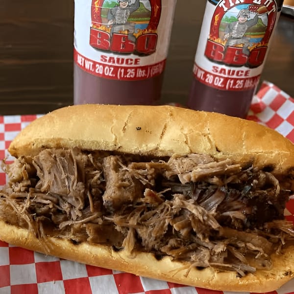 Tin Hut BBQ pulled pork sandwich on a red and white checkered paper with two sauce bottles.