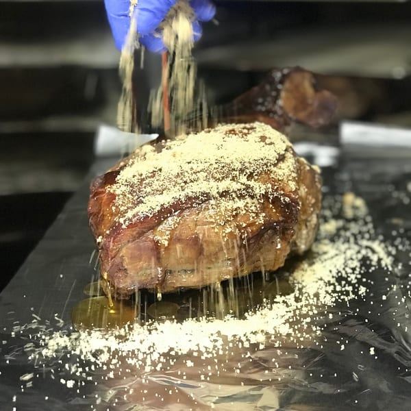 A gloved hand sprinkles seasoning over a large, glistening cut of Tin Hut BBQ meat.