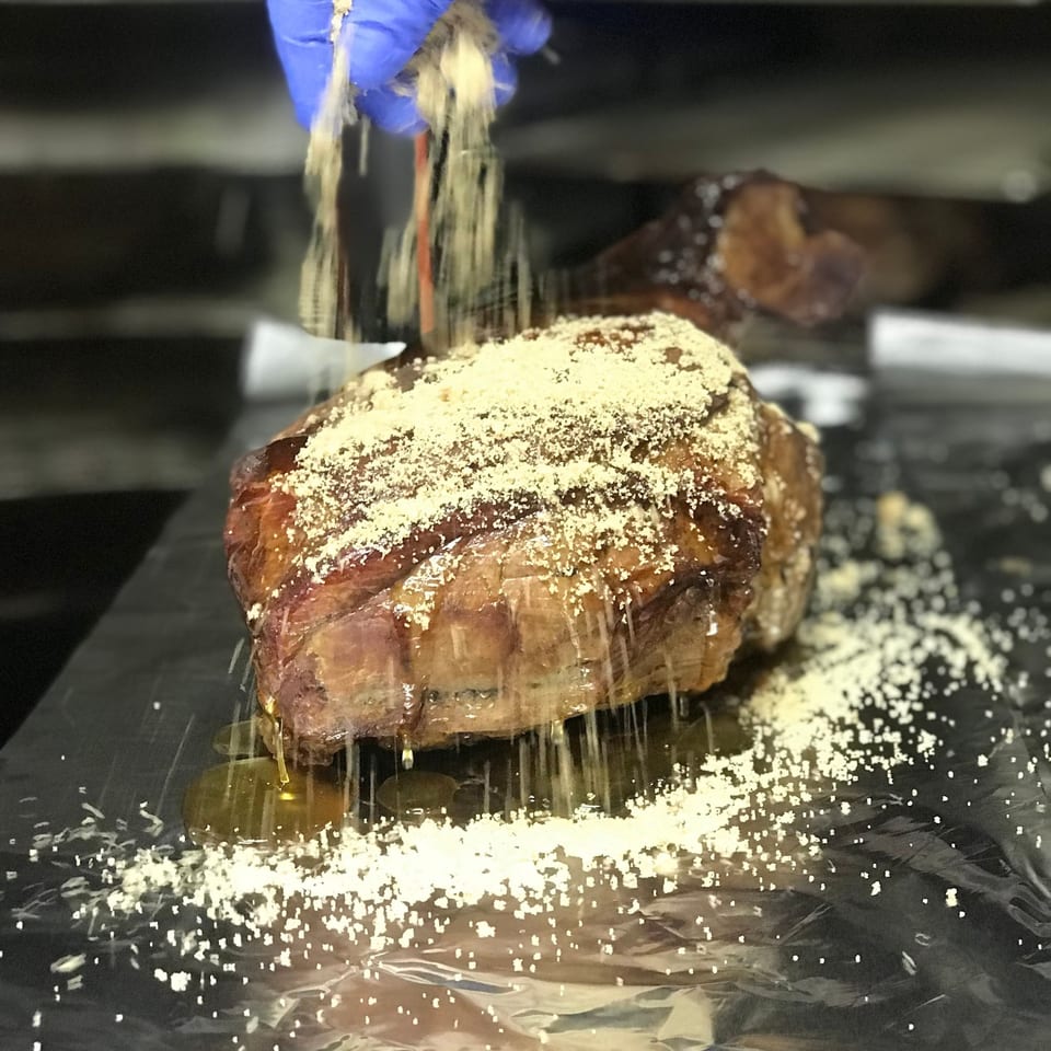 A gloved hand sprinkles seasoning over a large, glistening cut of Tin Hut BBQ meat.